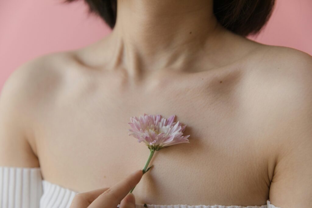 pexels photo 8182270 8182270 A close-up of a young woman holding a pink flower on her shoulder against a pastel pink background.